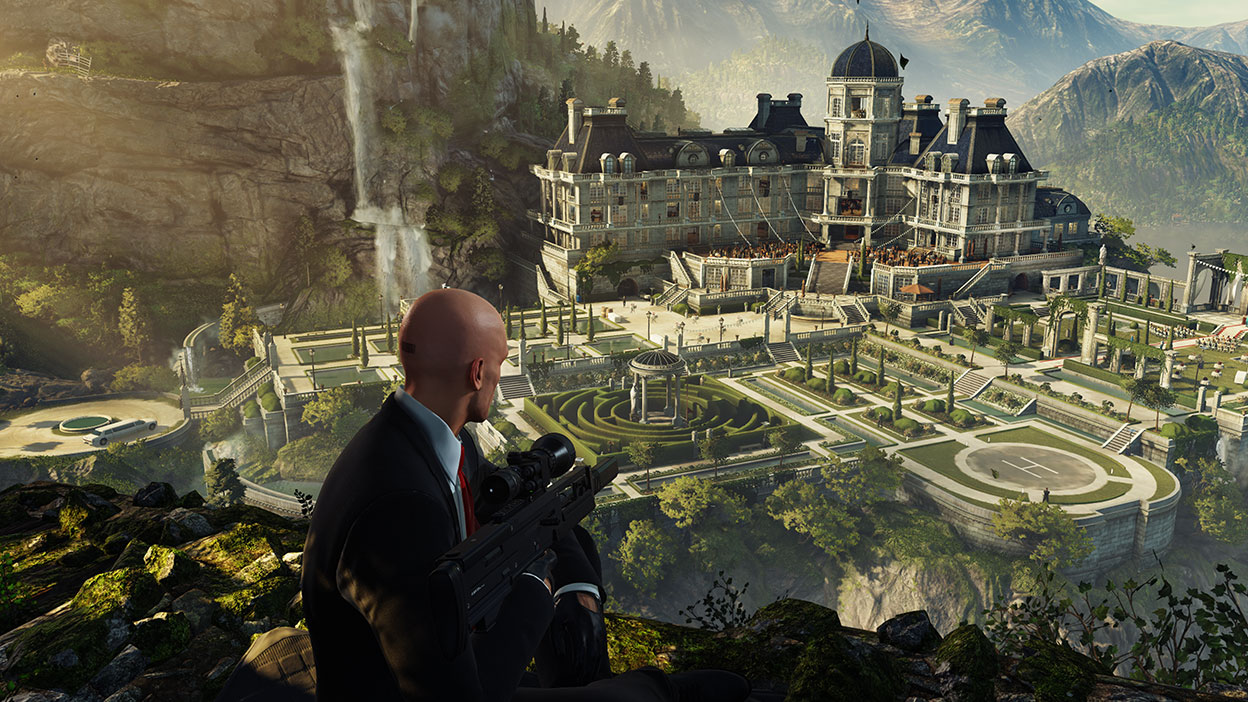 Agent 47 holds a sniper rifle while overlooking a large mansion on the side of a mountain
